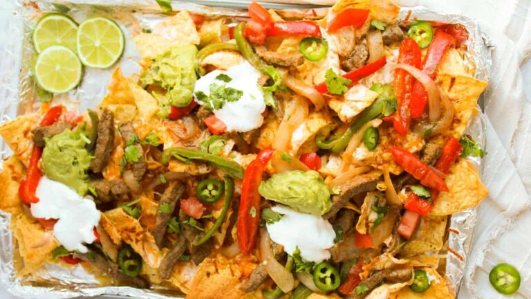 Sheet pan beef nachos topped with sliced steak, bell peppers, jalapenos, guacamole, sour cream, and fresh cilantro, served with lime slices on the side.