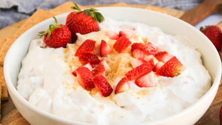 A white bowl filled with creamy dip topped with chopped fresh strawberries and a sprinkle of crumbs, with whole strawberries on the side.
