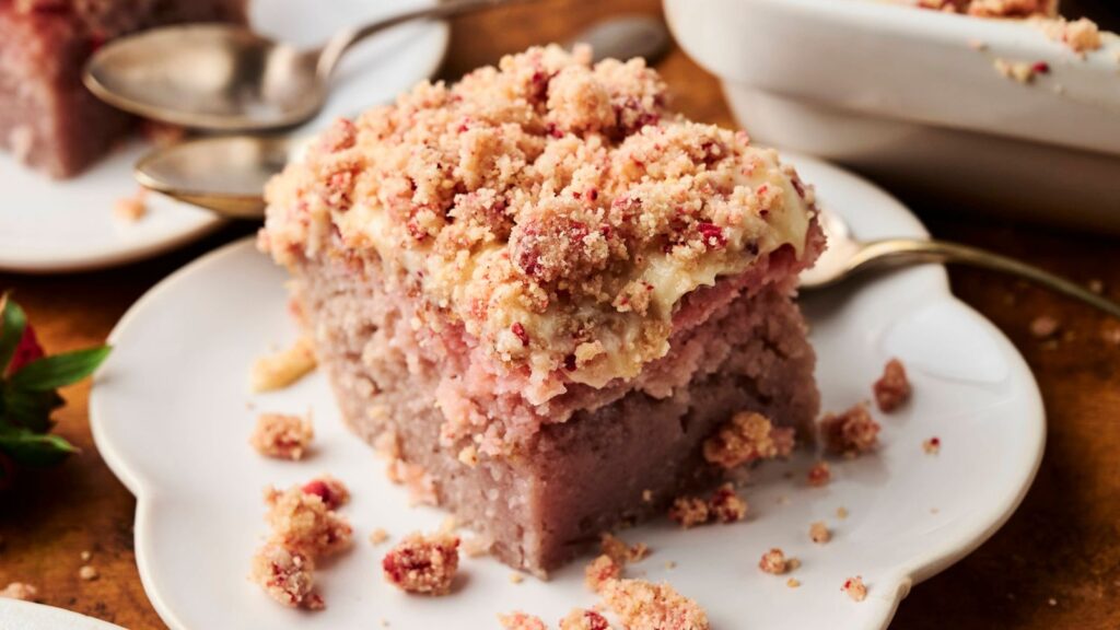 A square piece of crumb-topped cake with a pinkish hue sits on a white plate, with crumbs and a spoon nearby.