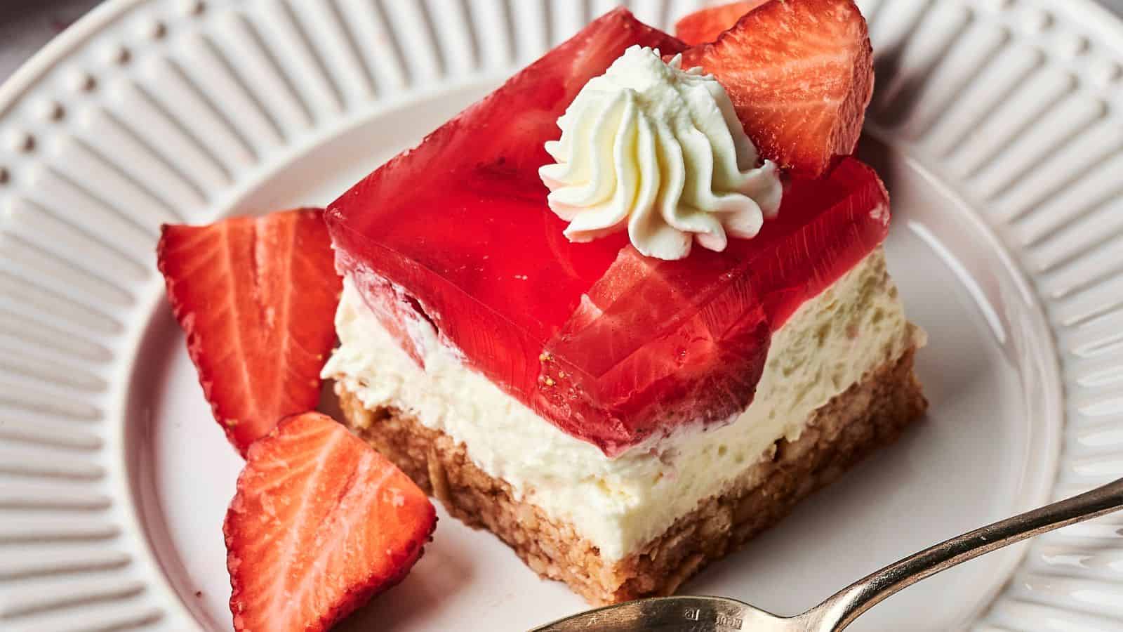 A dessert with a crumbly crust, creamy filling, a layer of red gelatin, topped with a whipped cream dollop and a strawberry slice, on a white plate with a spoon.