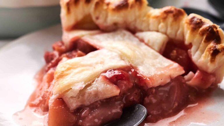 A close-up of a slice of fruit pie with a golden, flaky lattice crust and a juicy, mixed fruit filling on a plate.