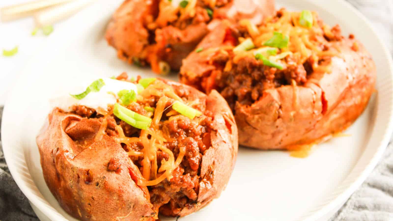 Three baked sweet potatoes topped with minced meat, melted cheese, sour cream, and chopped green onions on a white plate.