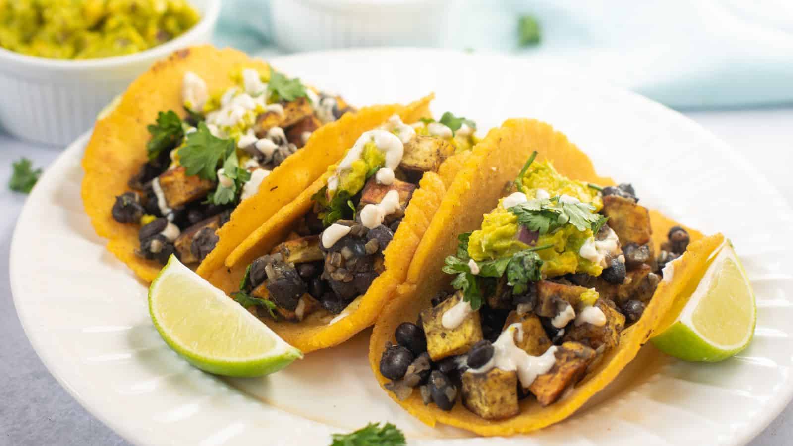 Three tacos filled with black beans, tofu, and vegetables, topped with sauce and cilantro, served with lime wedges on a white plate.