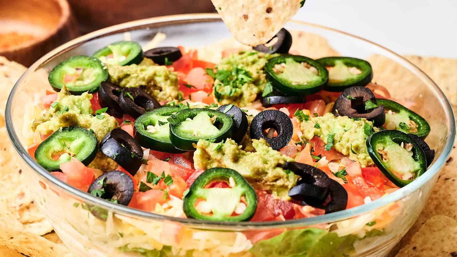 Glass bowl with layered dip topped with guacamole, sliced jalapeños, black olives, and tomatoes. A tortilla chip is dipping into the bowl.
