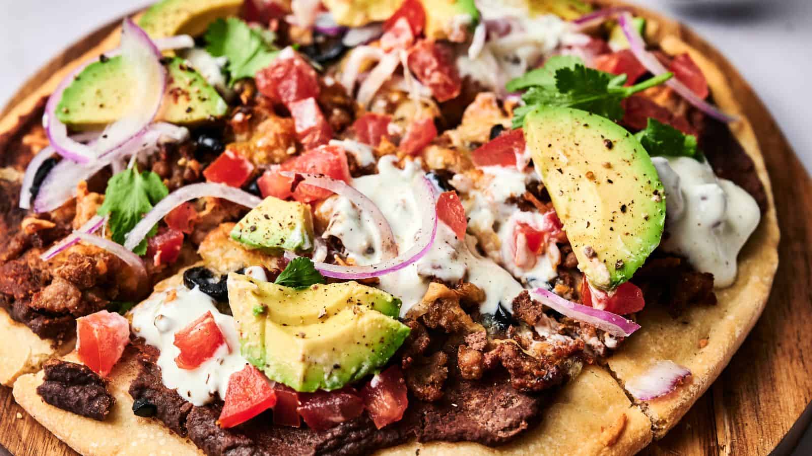 A close-up of a loaded flatbread pizza topped with avocado slices, diced tomatoes, onion, cilantro, seasoned meat, black beans, and a drizzle of creamy sauce.