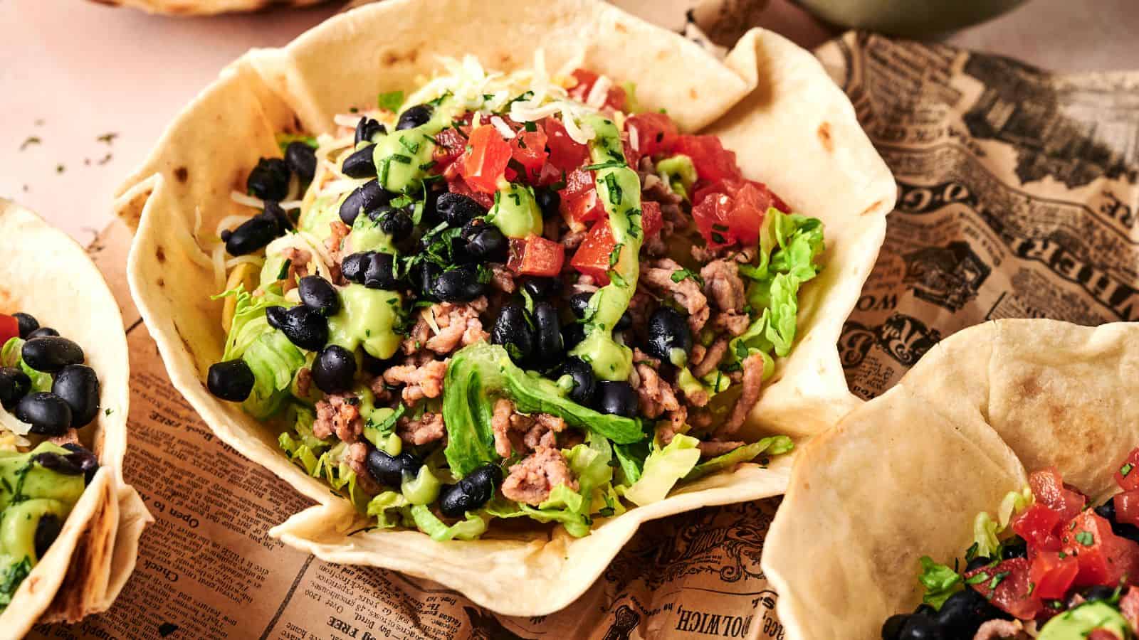 A taco salad served in an edible tortilla bowl, topped with ground meat, lettuce, black beans, diced tomatoes, avocado, and shredded cheese.