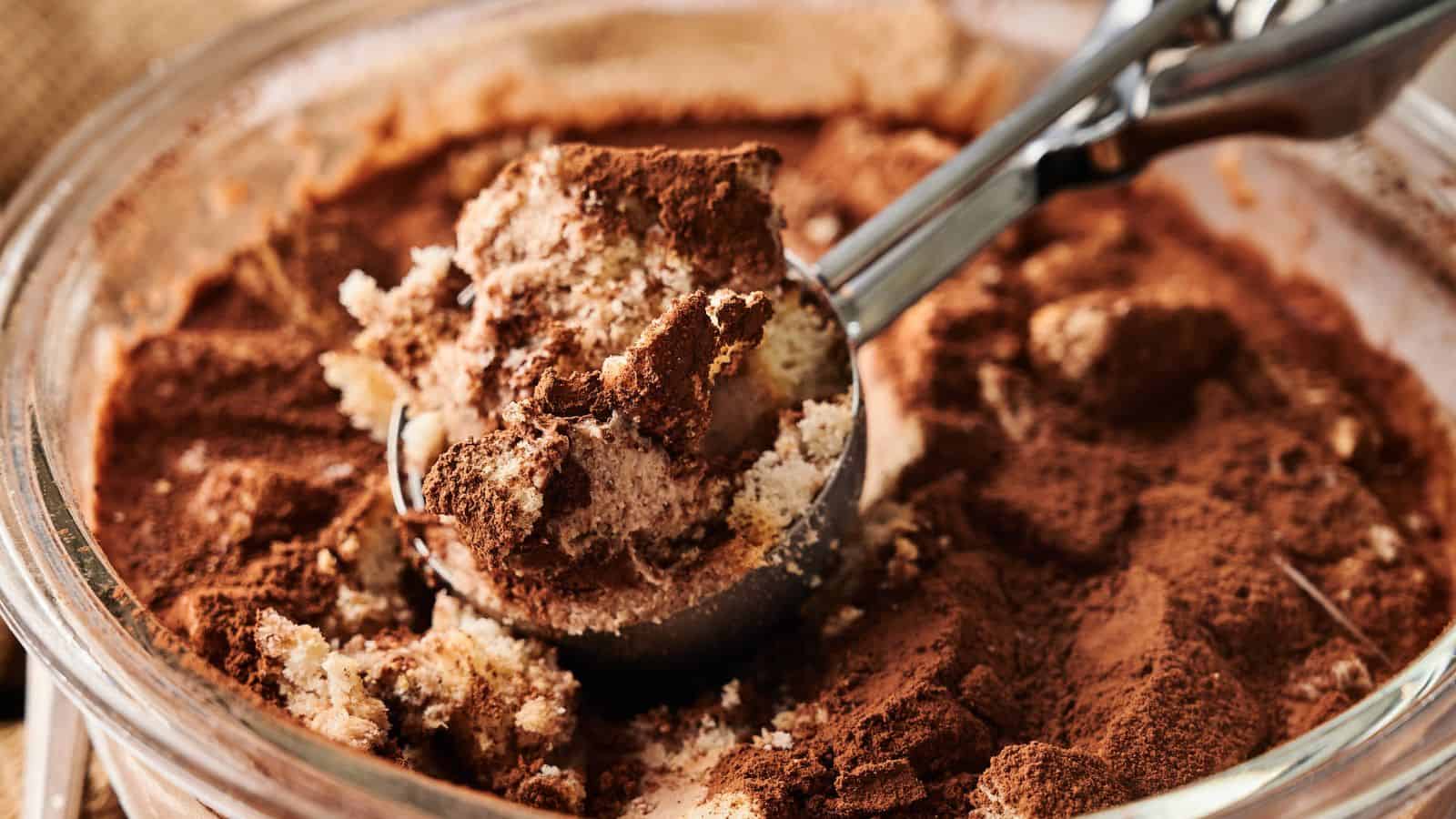 A close-up of a glass dish with tiramisu being scooped out, showing layers of cream, cake, and cocoa powder.