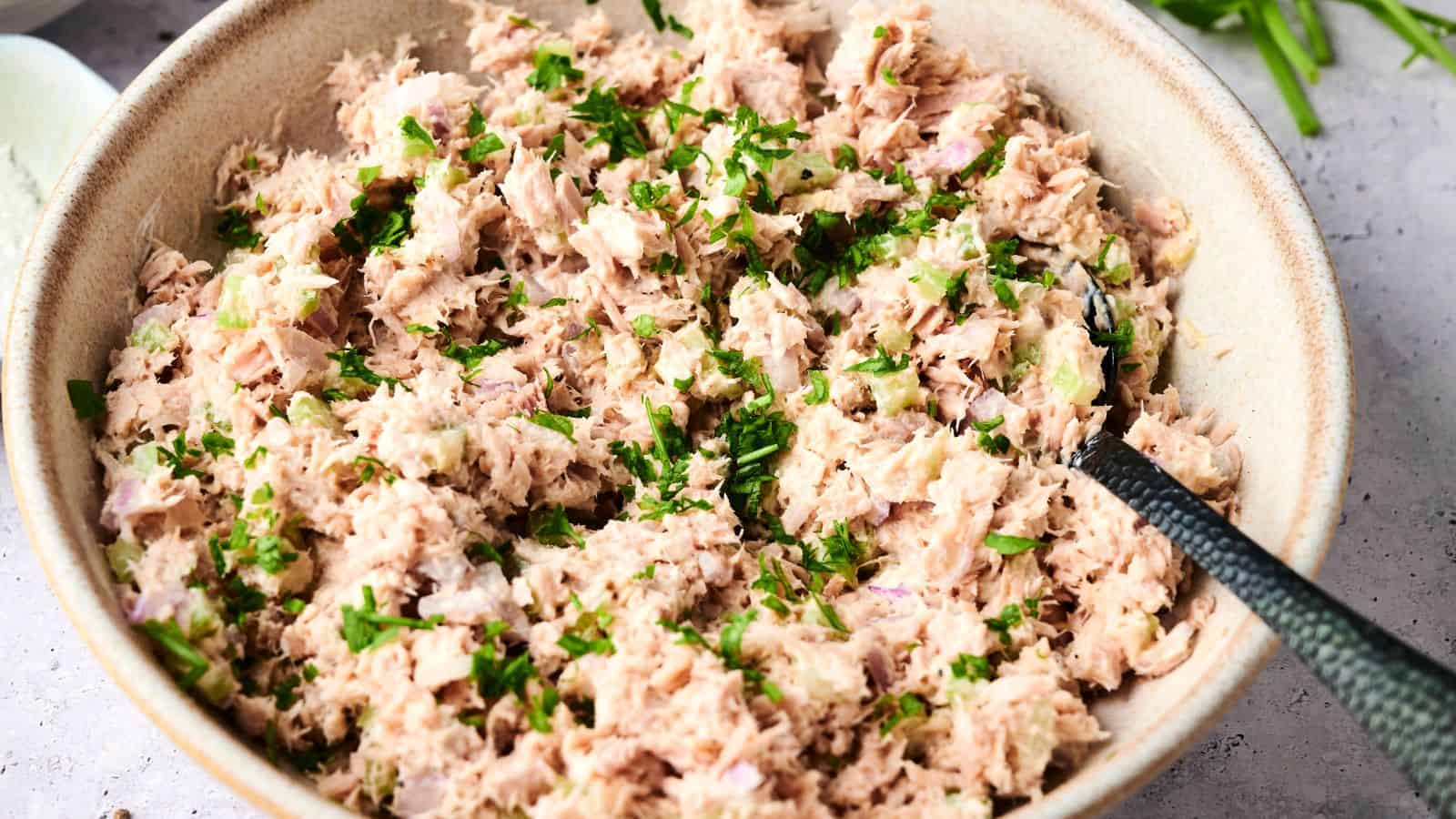 A bowl of tuna salad mixed with chopped herbs, likely parsley or chives, with a spoon resting inside the bowl.