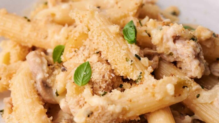Close-up of baked penne pasta with grated cheese, breadcrumbs, mushrooms, and fresh basil leaves.