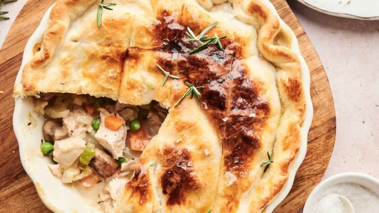 A golden-brown chicken pot pie with a slice removed, showing vegetables and chicken filling inside the crust, garnished with sprigs of rosemary.