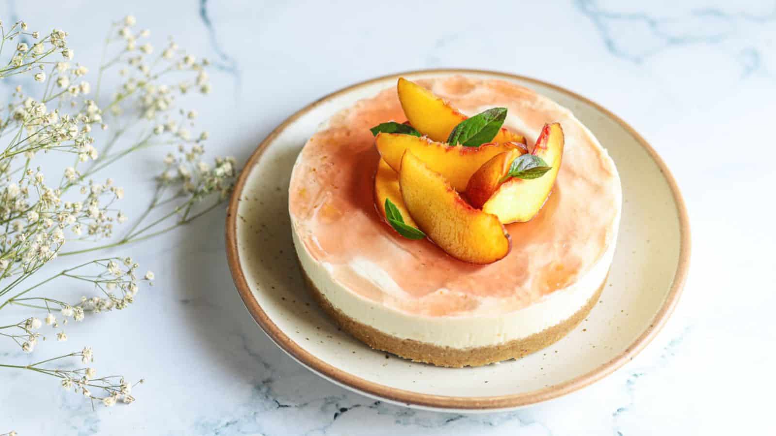 A peach-topped cheesecake on a beige plate, garnished with mint leaves, set on a light marble surface with sprigs of baby's breath flowers nearby.
