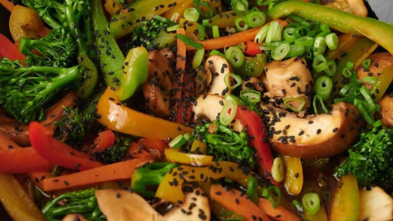 A close-up of a colorful vegetable stir-fry with mushrooms, bell peppers, broccoli, chopped scallions, and black sesame seeds.