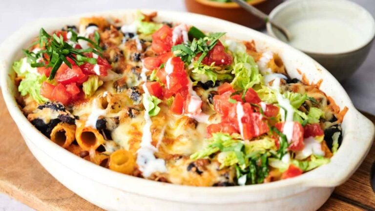 A baked pasta casserole topped with melted cheese, chopped lettuce, diced tomatoes, fresh herbs, and drizzled with white sauce in a white ceramic dish.