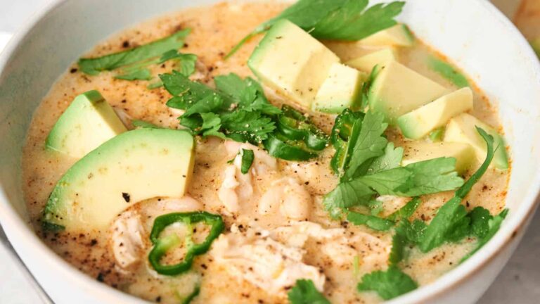 A bowl of creamy soup with shredded chicken, white beans, avocado slices, jalapeño rounds, and fresh cilantro garnished on top.
