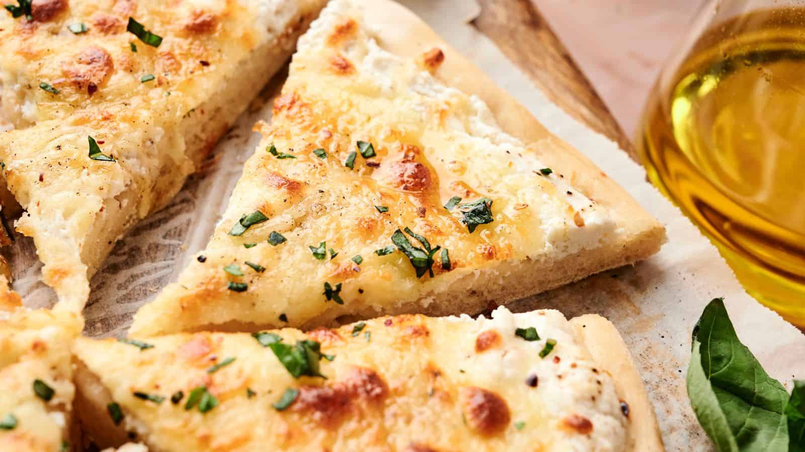 Close-up of sliced white pizza with melted cheese, fresh herbs, and a golden crust, served on parchment paper next to a glass container of olive oil.