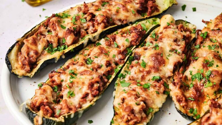 Four zucchini boats stuffed with ground meat, tomato sauce, and melted cheese, garnished with chopped parsley, served on a white plate.
