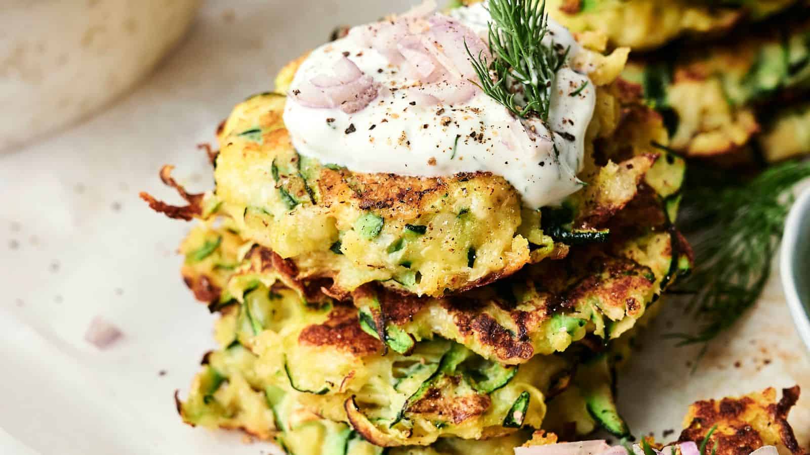 A stack of zucchini fritters topped with sour cream, thinly sliced shallots, and a sprig of dill.