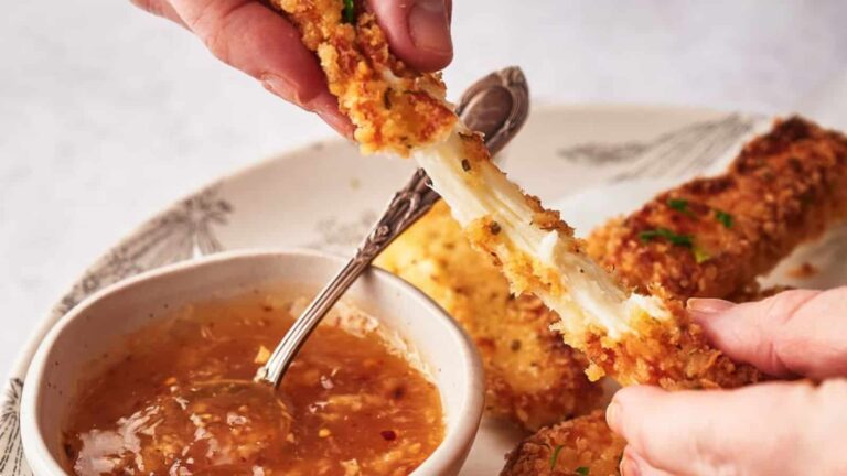 Hands pull apart a breaded mozzarella stick with melted cheese, next to a bowl of dipping sauce on a plate.
