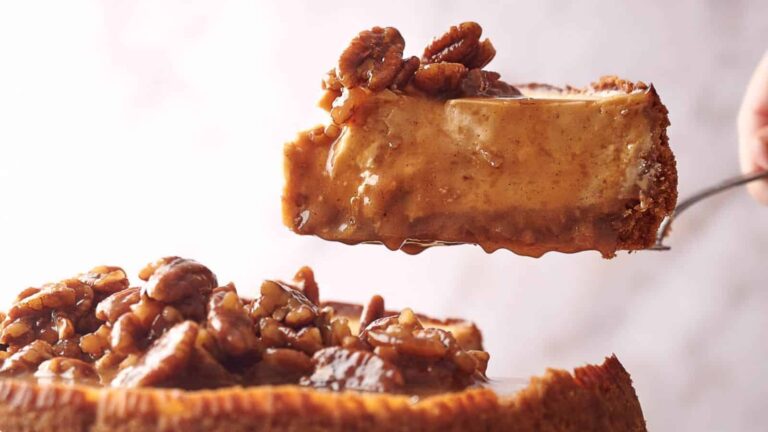 A close-up of a slice of cheesecake topped with glazed pecans being lifted from the whole cake.