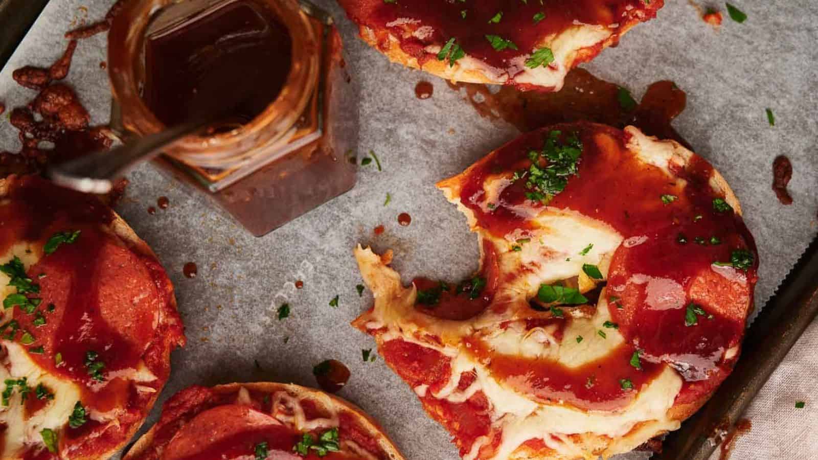 Pizza bagels on parchment paper, one with a bite out of it and a jar of BBQ sauce nearby.