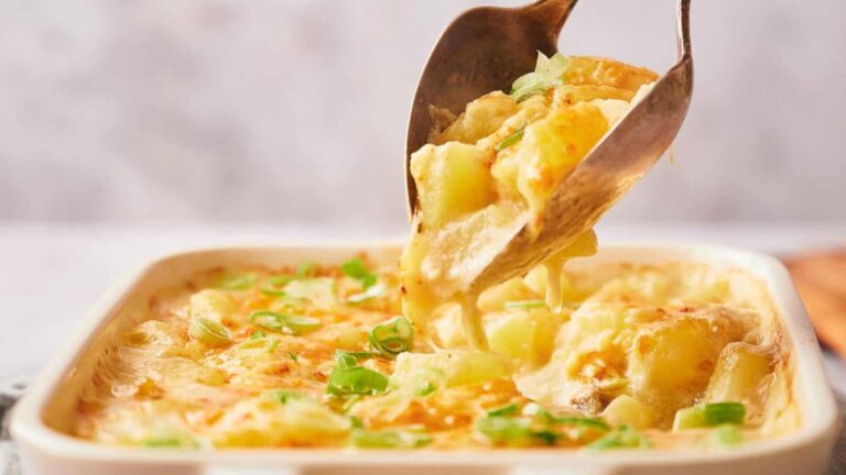 A serving spoon lifts a portion of cheesy potato casserole with green onions from a baking dish.