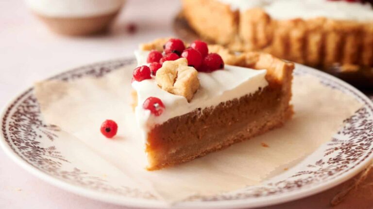 A slice of pie with a creamy white topping, garnished with red berries, sits on a patterned plate with a larger pie in the background.