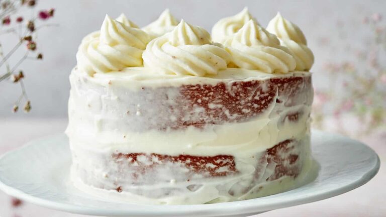 A partially frosted layer cake with white icing and piped swirls on top, displayed on a white plate.