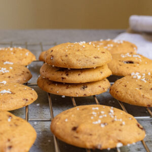 A stack of Salted Honey Cookies topped with coarse salt sits on a cooling rack, with more cookies and a towel in the background.