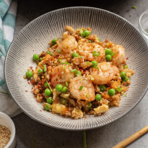 A bowl of Shrimp Cauliflower Fried Rice with peas, scrambled eggs, and vegetables sits on a gray surface, accompanied by chopsticks and a skillet in the background.