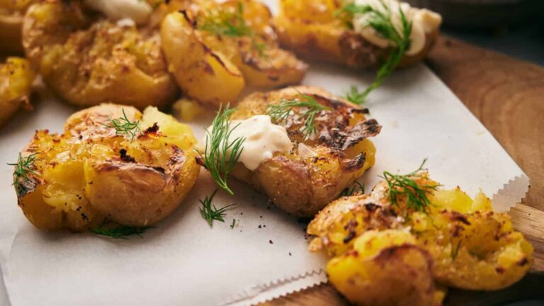 Crispy smashed potatoes topped with fresh dill and a dollop of creamy sauce, served on parchment paper.