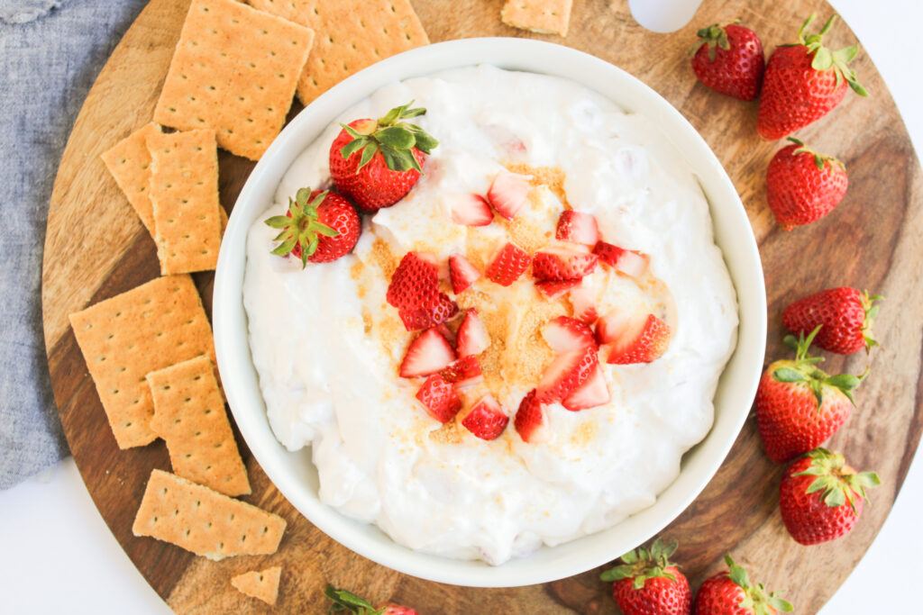 A bowl of creamy Strawberry Cheesecake Dip topped with chopped strawberries and crumbs, surrounded by whole strawberries and graham crackers on a wooden board.