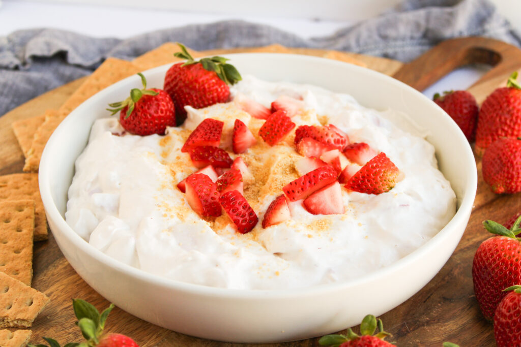 A white bowl filled with creamy Strawberry Cheesecake Dip, topped with chopped strawberries and graham cracker crumbs, is surrounded by fresh strawberries and graham crackers on a wooden board.