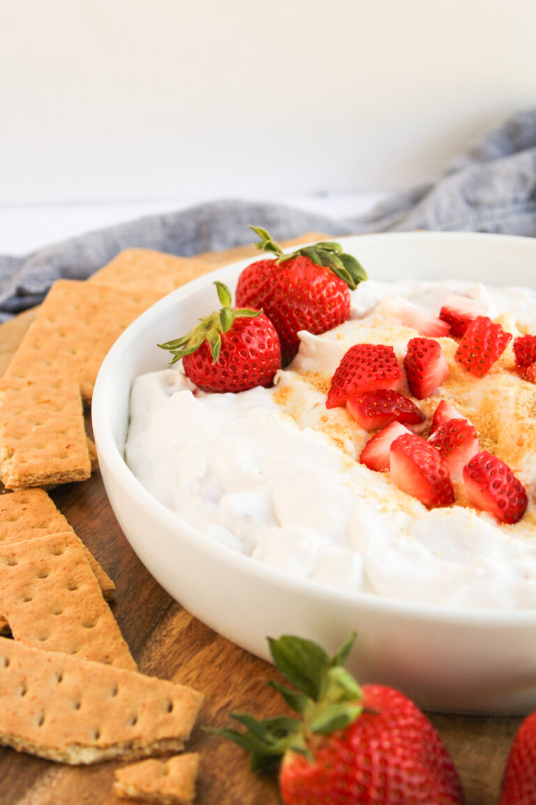 A bowl of creamy Strawberry Cheesecake Dip topped with sliced strawberries and graham cracker crumbs, surrounded by whole strawberries and graham crackers on a wooden surface.