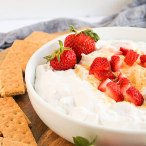A bowl of creamy Strawberry Cheesecake Dip topped with sliced strawberries and graham cracker crumbs, surrounded by whole strawberries and graham crackers on a wooden surface.