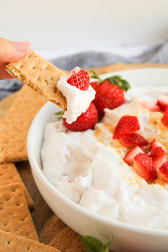A hand dips a piece of graham cracker into a bowl of strawberry cheesecake dip, topped with fresh strawberries.