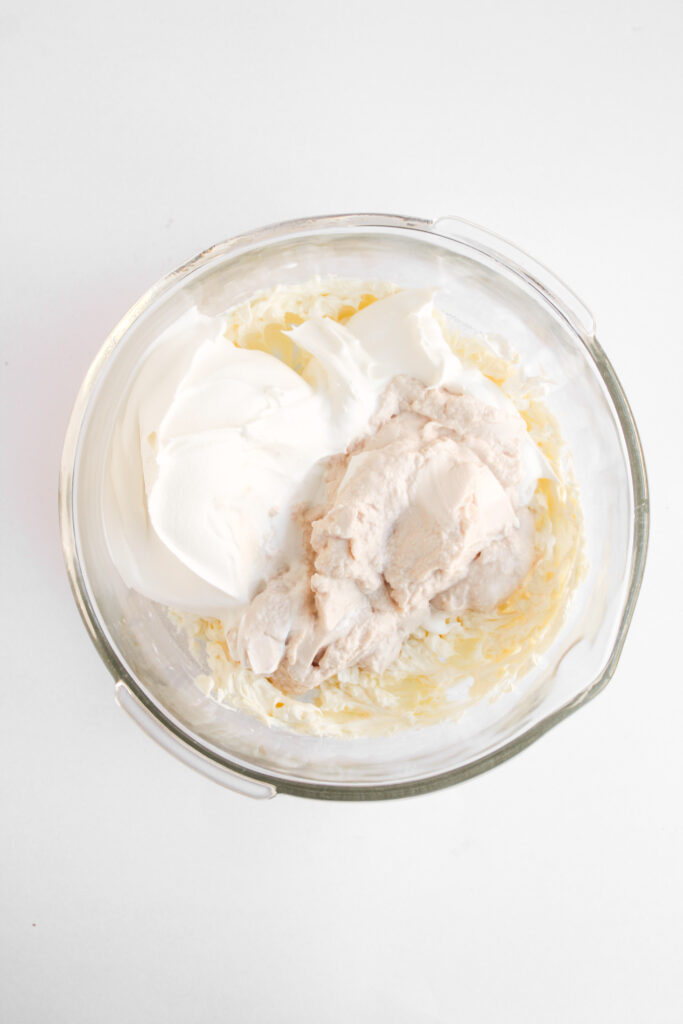 A glass bowl containing a mixture of whipped cream cheese, sour cream, and a thick beige substance sits on a white surface&mdash;perfect for enjoying as a creamy Strawberry Cheesecake Dip.