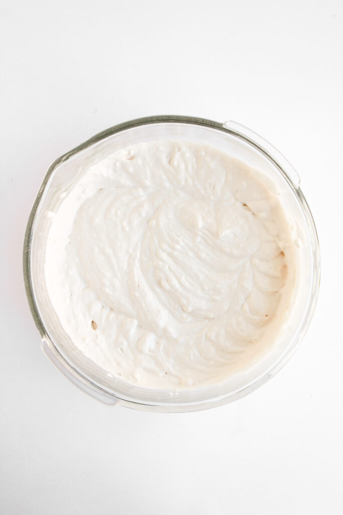A glass bowl filled with a creamy white mixture, possibly Strawberry Cheesecake Dip or batter, on a white background.