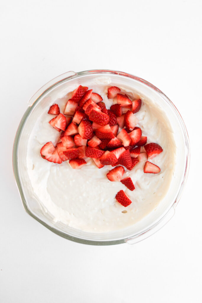 A glass bowl filled with creamy white Strawberry Cheesecake Dip, topped with chopped fresh strawberries and placed on a white surface.