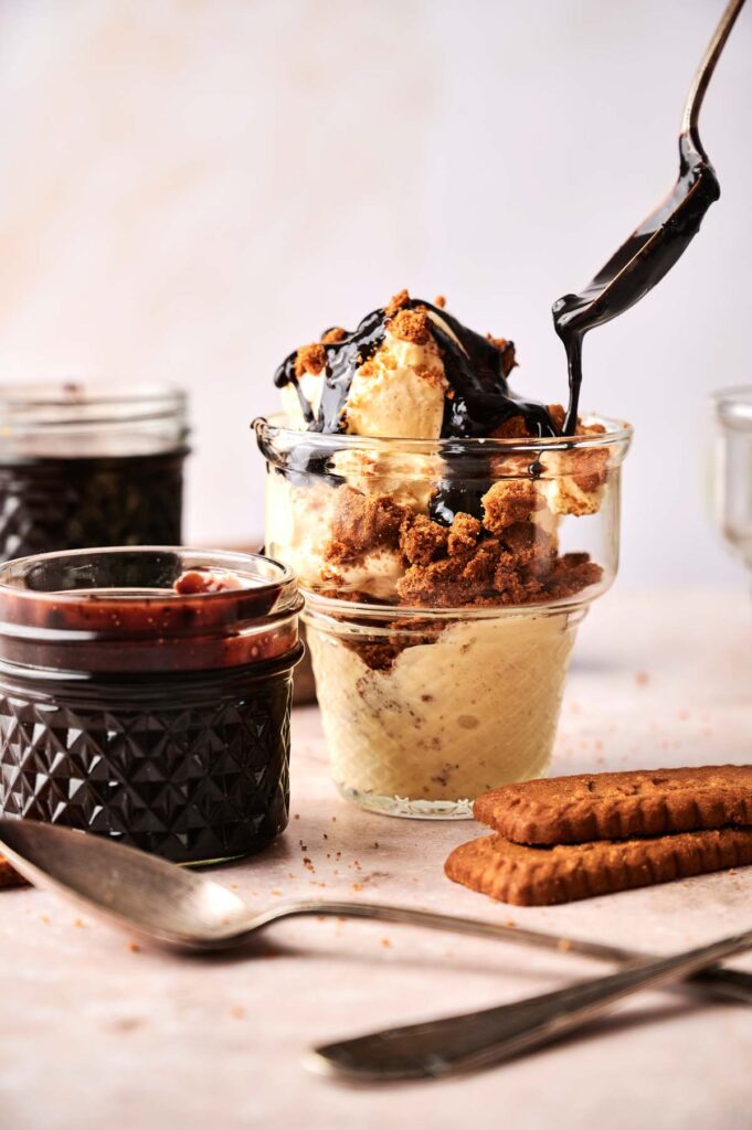 A glass of vanilla ice cream topped with cookie crumbs and chocolate sauce creates the perfect Hot Fudge Sundae, surrounded by jars of sauce and cookies on a table.