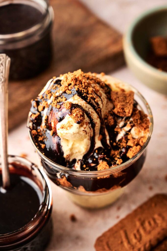 A glass cup of vanilla ice cream topped with rich hot fudge sundae sauce and crumbled cookies, with a jar of chocolate sauce and a cookie nearby.