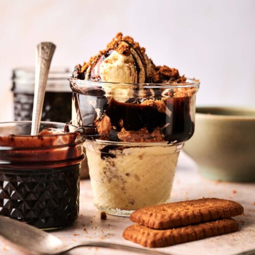 A glass dessert cup filled with vanilla ice cream, rich chocolate sauce, and crumbled cookies creates a Hot Fudge Sundae vibe, with jars of sauce and whole cookies on the table.