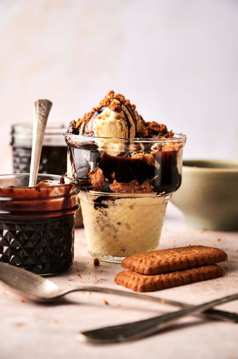 A glass dessert cup filled with vanilla ice cream, rich chocolate sauce, and crumbled cookies creates a Hot Fudge Sundae vibe, with jars of sauce and whole cookies on the table.