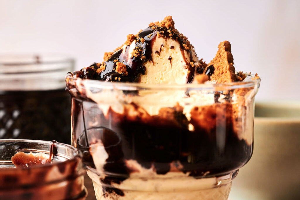A glass filled with vanilla ice cream topped with rich chocolate sauce and crumbled cookies, creating a classic Hot Fudge Sundae, with jars in the background.