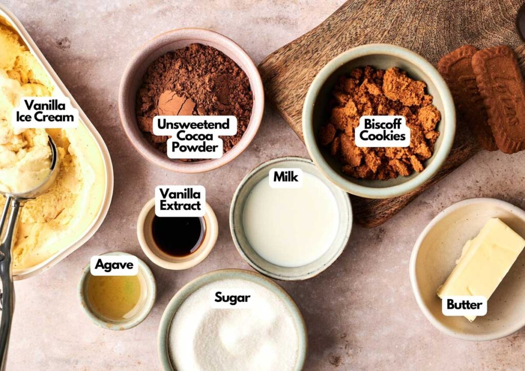 Several bowls and dishes containing vanilla ice cream, unsweetened cocoa powder, Biscoff cookies, milk, butter, agave, sugar, and vanilla extract are arranged on a countertop for assembling a delicious Hot Fudge Sundae.