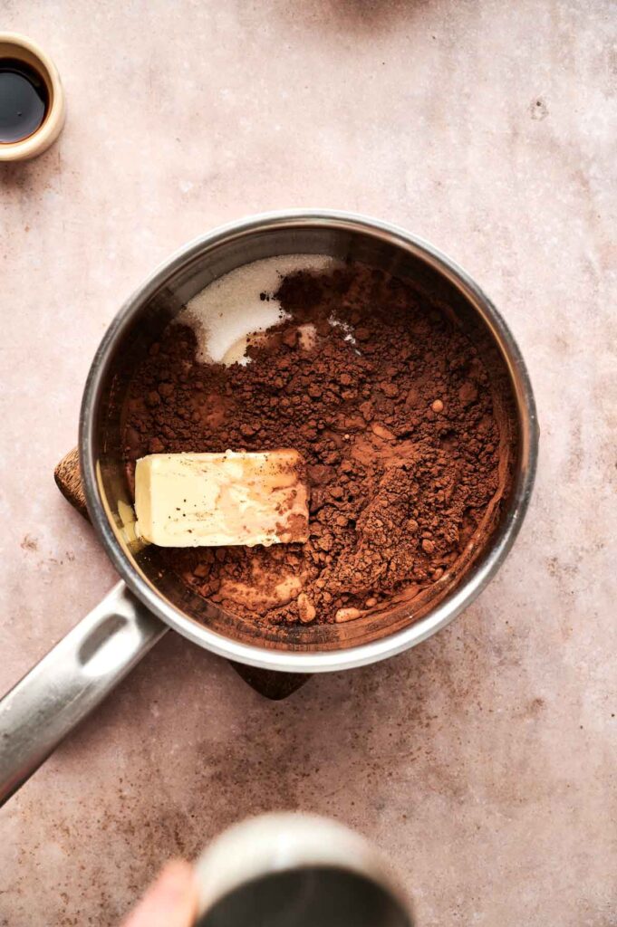 A saucepan containing cocoa powder and a slab of butter, perfect for making Hot Fudge Sundae sauce, sits on a light countertop, viewed from above.
