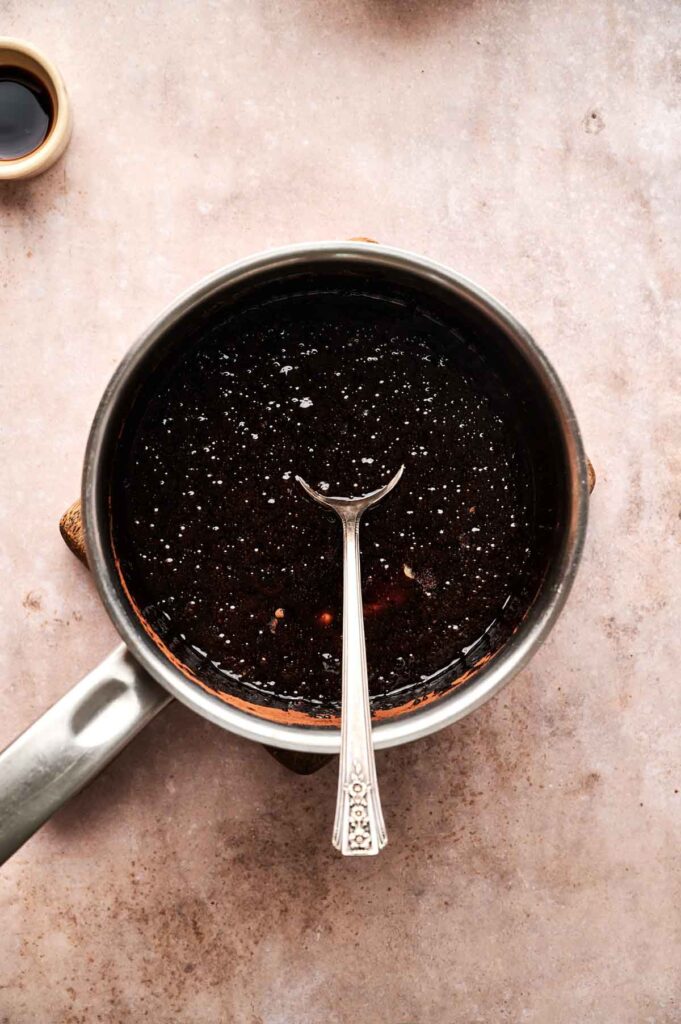 A saucepan filled with a dark liquid, possibly hot fudge for a sundae, with a spoon resting inside on a light brown countertop.
