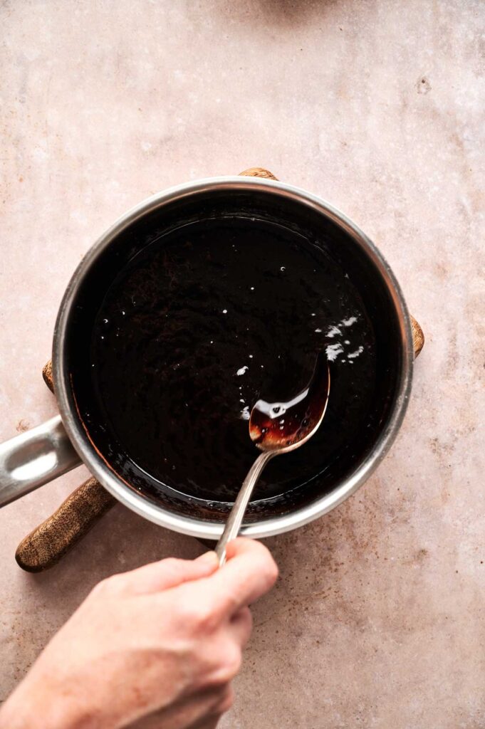 A hand holding a spoon stirs a dark liquid—like the beginnings of a Hot Fudge Sundae sauce—in a saucepan on a light-colored countertop.