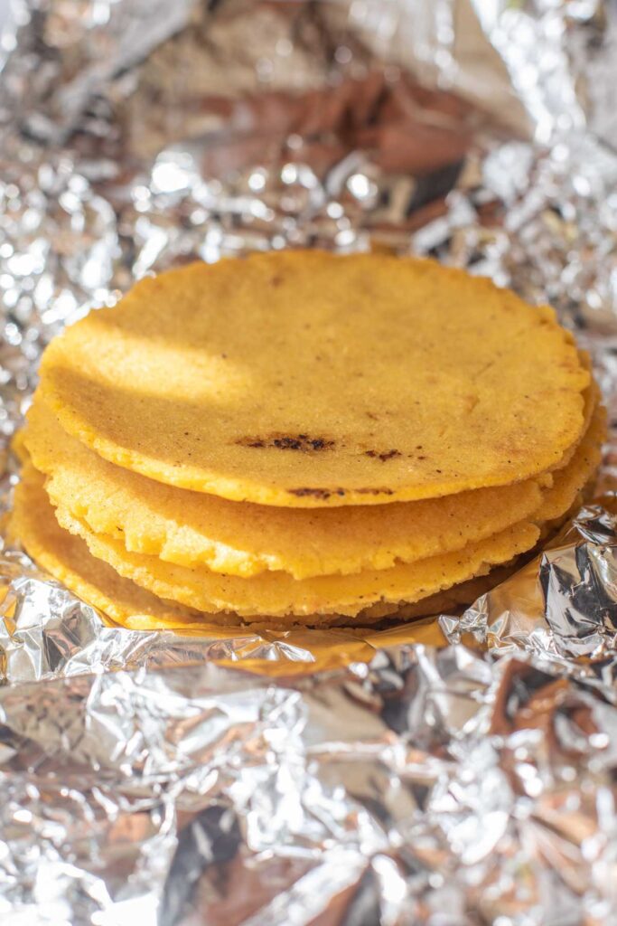 A stack of corn tortillas rests on crumpled aluminum foil, with sunlight highlighting their yellow color and texture&mdash;perfect for assembling Sweet Potato and Black Bean Tacos.