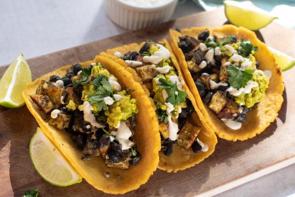 Three Sweet Potato and Black Bean Tacos filled with diced tofu, guacamole, cilantro, and drizzled sauce on a wooden board, served with fresh lime wedges.