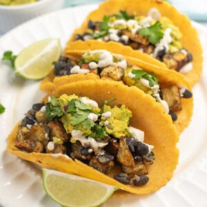 Three Sweet Potato and Black Bean Tacos filled with grilled vegetables, guacamole, cheese, and cilantro are served on a white plate with lime wedges.